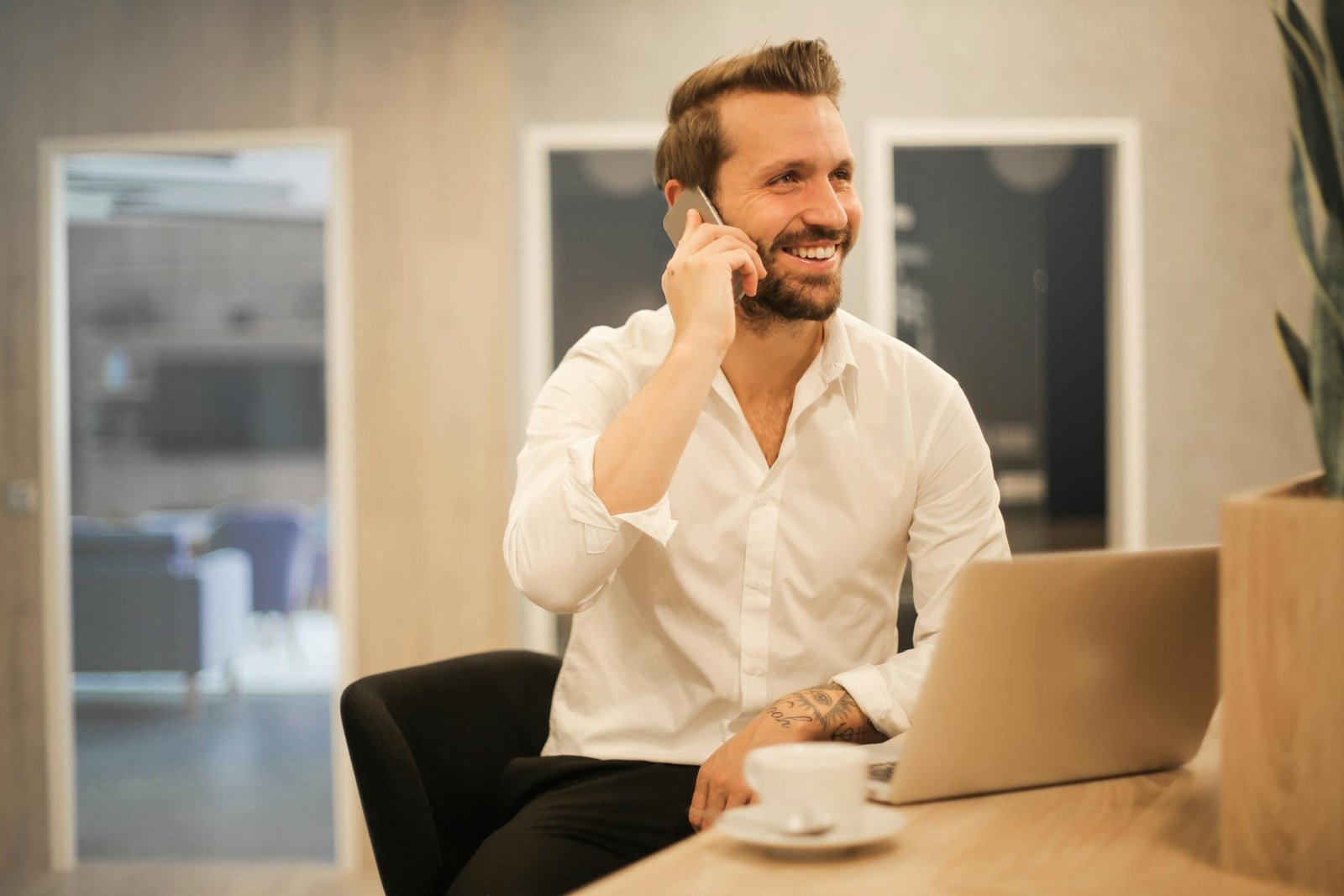 Happy businessman in white shirt on phone call at modern office desk with laptop.